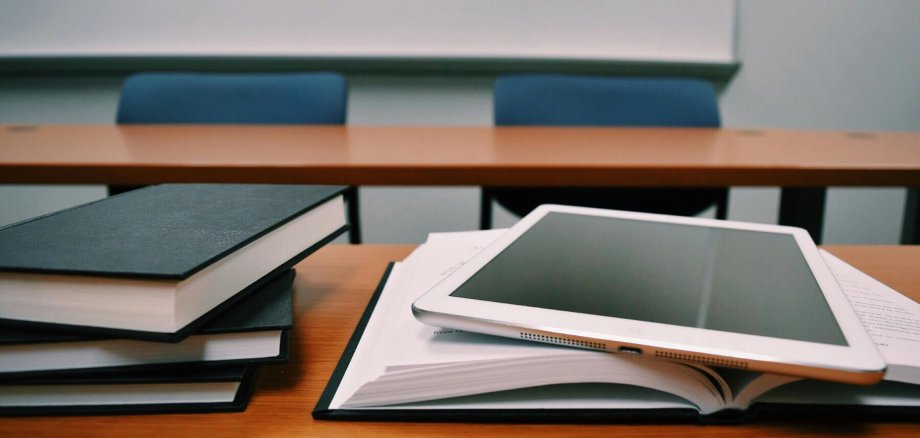 Ein Tablet liegt auf einem geöffneten Buch auf einem Schreibtisch. Daneben sind mehrere gestapelte Bücher zu sehen. Im Hintergrund stehen zwei leere Stühle vor einem weiteren Tisch. Der Raum wirkt wie ein Klassenzimmer oder Seminarraum.