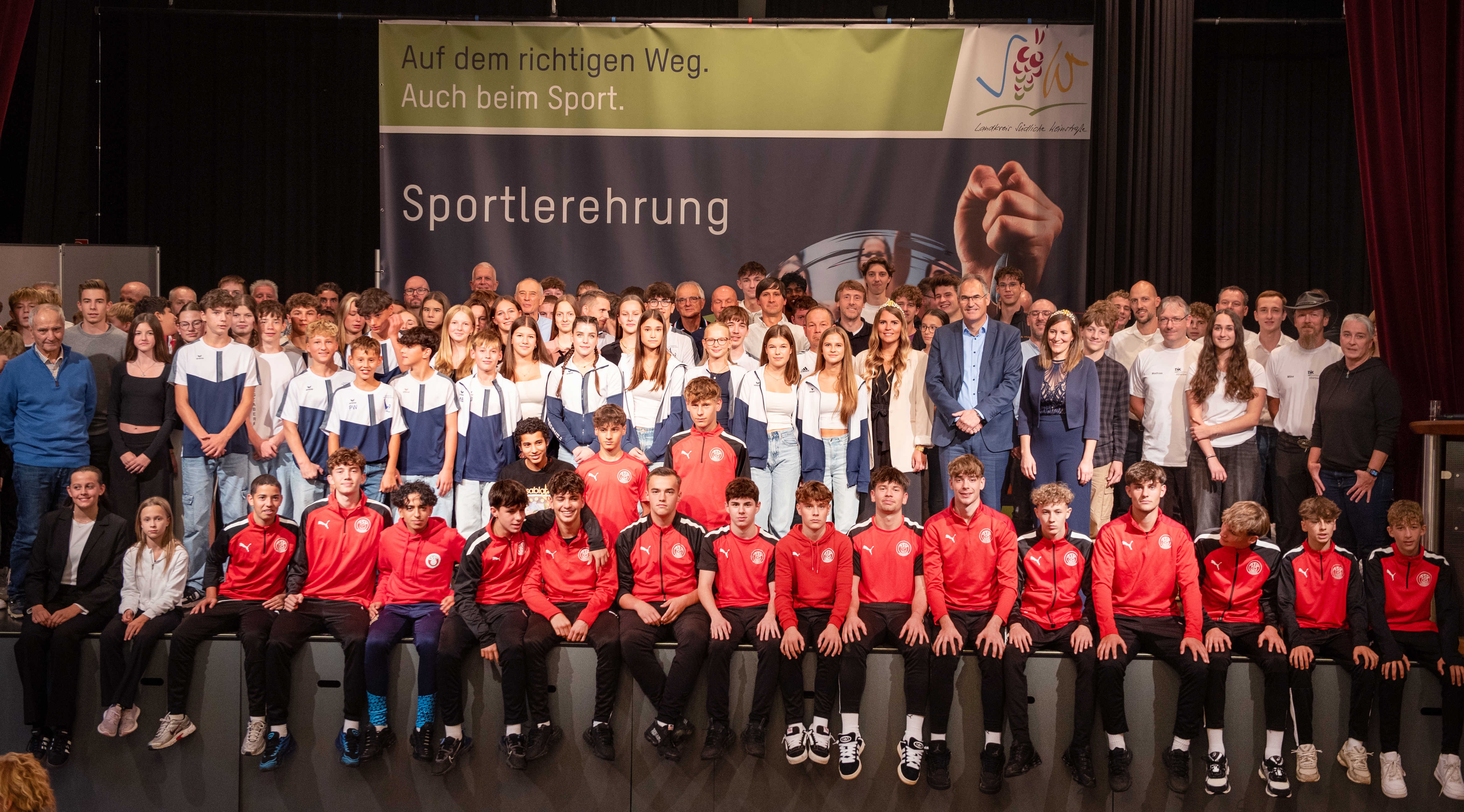 Eine große Gruppe von Sportlerinnen und Sportlern steht dicht beieinander auf der Bühne des Hohenstaufensaals in Annweiler. Vor einem Banner mit der Aufschrift "Sportlerehrung" posieren die Geehrten für ein Gruppenfoto. Im Hintergrund ist das Motto "Auf dem richtigen Weg. Auch beim Sport." zu lesen. Zwischen den Sportlern steht auch Landrat Dietmar Seefeldt.