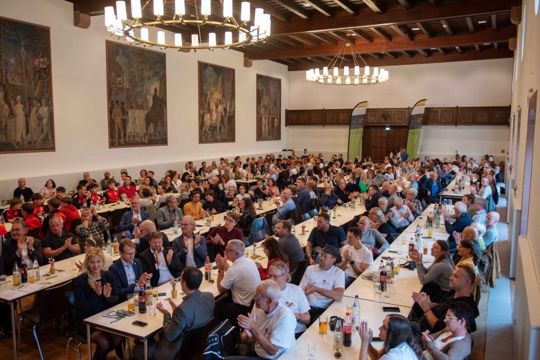 Viel Applaus gab es im Hohenstaufensaal für die sportlichen Erfolge. Foto: Iversen Der Hohenstaufensaal in Annweiler ist gefüllt mit rund 300 Gästen, die applaudierend an Tischen sitzen. Die Wände des Saals sind mit großen Gemälden verziert, und von der Decke hängen Kronleuchter. Die Gäste würdigen die sportlichen Erfolge der Geehrten.