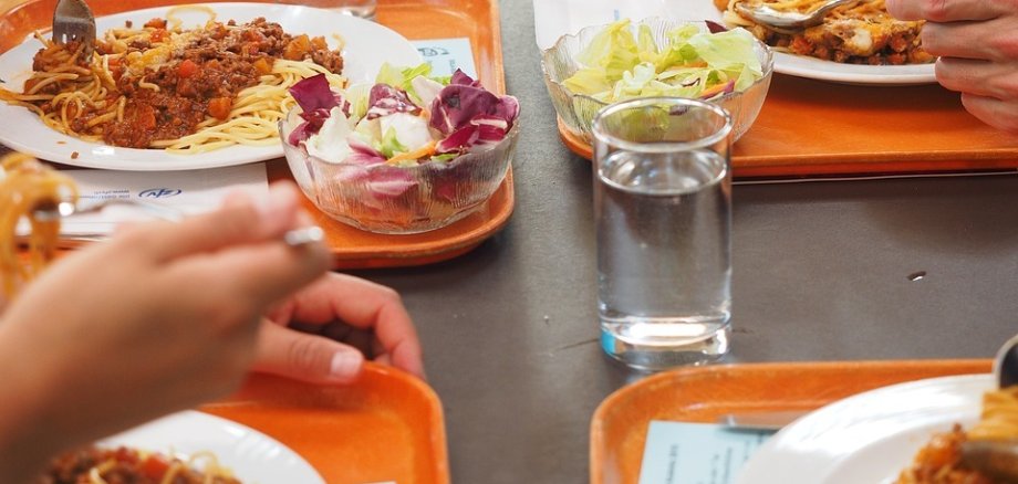 Nahaufnahme eines Tischs in einer Kantine, an dem mehrere Personen sitzen und Spaghetti mit Bolognese-Soße essen. Auf den Tabletts befinden sich auch Gläser mit Wasser und kleine Schüsseln mit gemischtem Salat. Die Hände der Personen sind mit Besteck beschäftigt.