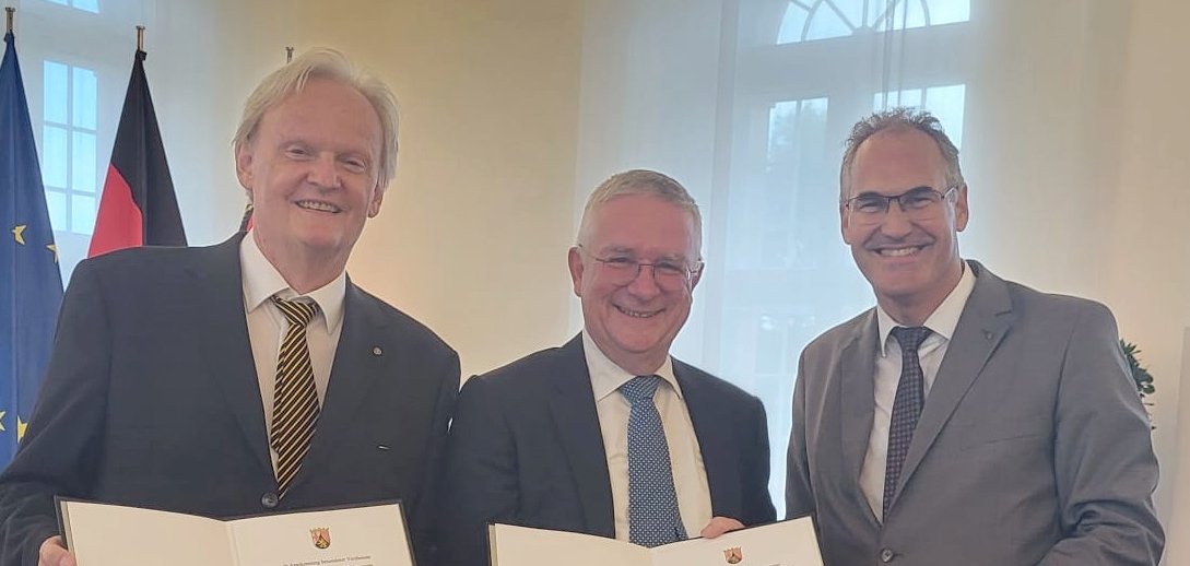 Auf dem Bild sind drei Männer in Anzügen zu sehen, die jeweils eine Urkunde in den Händen halten und lächeln. Sie stehen in einem festlichen Raum mit einem Kronleuchter an der Decke. Im Hintergrund sind die Flaggen der Europäischen Union und Deutschlands zu erkennen. Rechts im Bild steht Landrat Dietmar Seefeldt, in der Mitte Hansjörg Rebholz und links Dr. Volker Thorn. Beide Männer in der Mitte und links wurden für ihr herausragendes berufsbezogenes Engagement ausgezeichnet.