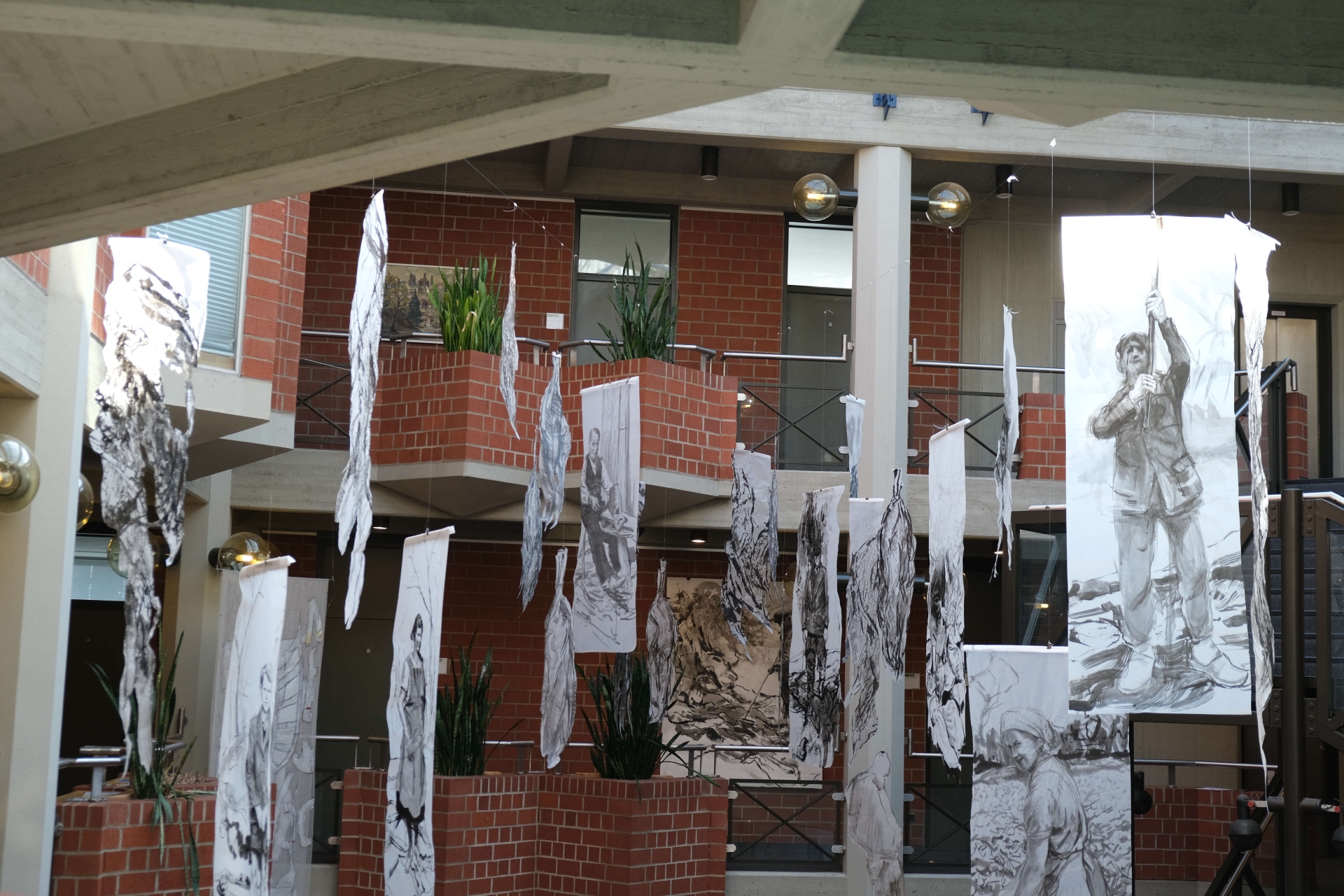 Die preisgekrönte Installation „Tabak“ (2024) steht im Mittelpunkt der Schau. Foto: KV SÜW Kunstinstallation im Kreishaus Südliche Weinstraße, einem offenen, mehrstöckigen Gebäude mit Backsteinwänden und großen Fenstern. Mehrere längliche Bahnen mit schwarz-weißen Zeichnungen von Menschen und Pflanzen hängen von der Decke herab. Die Motive zeigen unter anderem Männer und Frauen bei landwirtschaftlicher Arbeit.