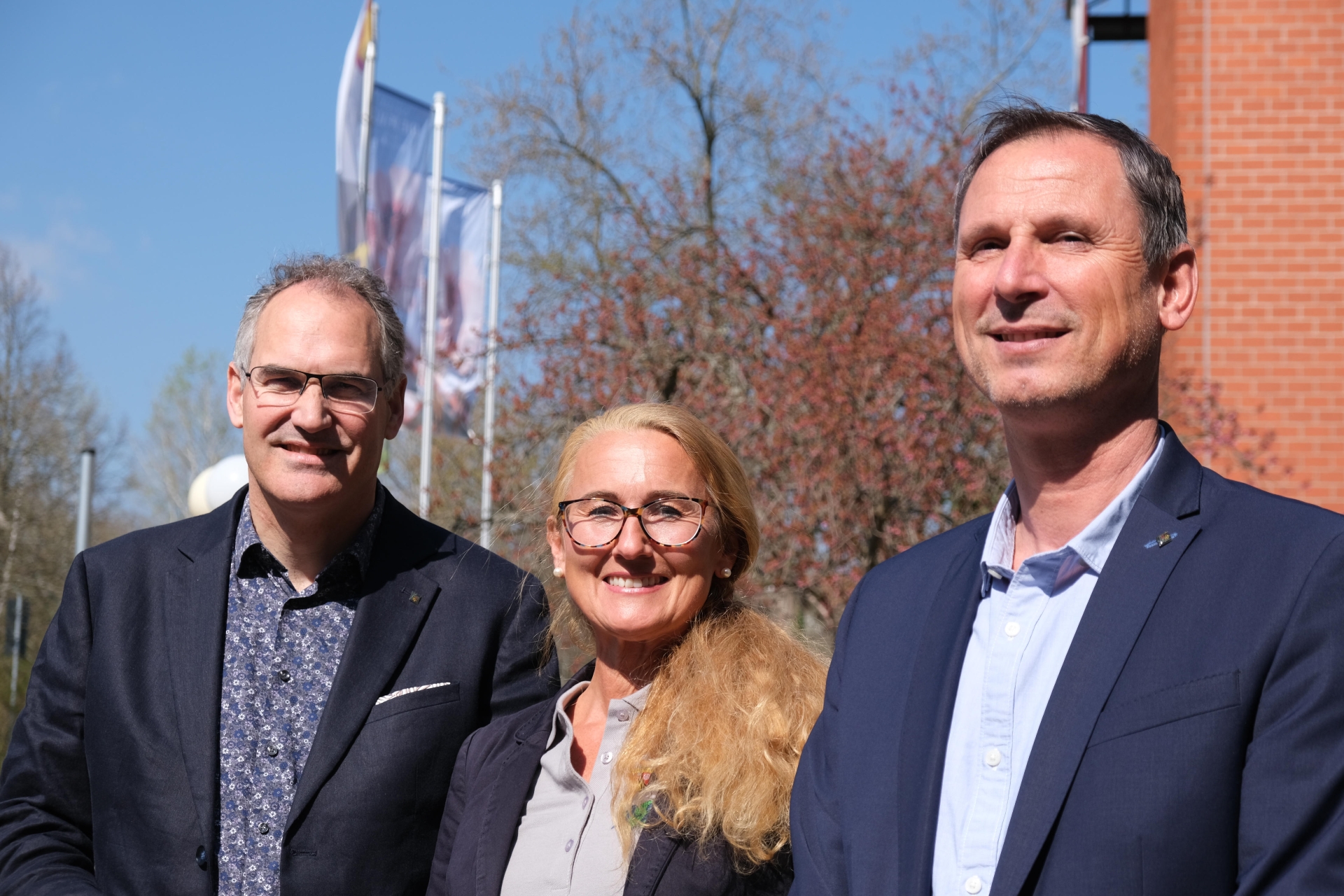 Landrat Dietmar Seefeldt, Bewegungsmanagerin Vera Krupinski und Sportreferent Heiko Pabst im Porträt vor dem Kreishaus bei blauem Himmel und Sonnenschein.