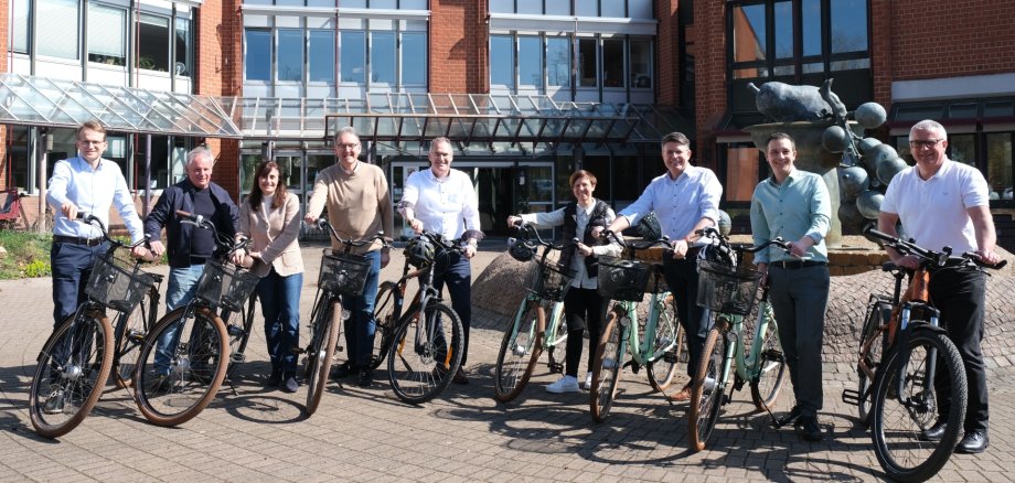 Werben gemeinsam fürs STADTRADELN 2025, von links nach rechts: Daniel Salm, Bürgermeister der Verbandsgemeinde Edenkoben, Werner Schreiner (Kreisbeigeordneter), Gabriele Flach (Bürgermeisterin der Verbandsgemeinde Maikammer), Georg Kern (Erster Kreisbeigeordneter), Landrat Dietmar Seefeldt, Kathrin Flory (Bürgermeisterin Verbandsgemeinde Bad Bergzabern), Torsten Blank (Bürgermeister der Verbandsgemeinde Landau-Land), Christian Burkhart (Bürgermeister der Verbandsgemeinde Annweiler am Trifels) und Christian Sommer (Bürgermeister der Verbandsgemeinde Herxheim). Foto: KV SÜW Neun Personen stehen nebeneinander mit Fahrrädern vor dem Verwaltungsgebäude der Kreisverwaltung Südliche Weinstraße. Alle lächeln in die Kamera und werben gemeinsam für das STADTRADELN 2025. Im Hintergrund ist ein rotes Ziegelgebäude mit Glasfassade und eine Brunnen-Skulptur zu sehen.