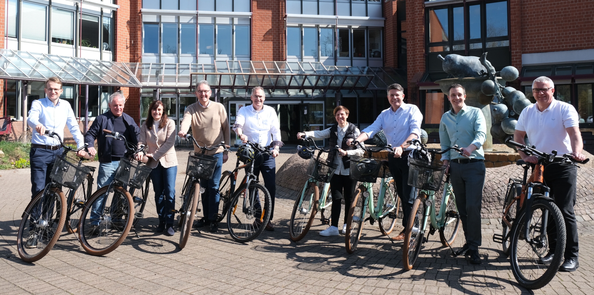 Neun Personen stehen nebeneinander mit Fahrrädern vor dem Verwaltungsgebäude der Kreisverwaltung Südliche Weinstraße. Alle lächeln in die Kamera und werben gemeinsam für das STADTRADELN 2025. Im Hintergrund ist ein rotes Ziegelgebäude mit Glasfassade und eine Brunnen-Skulptur zu sehen.