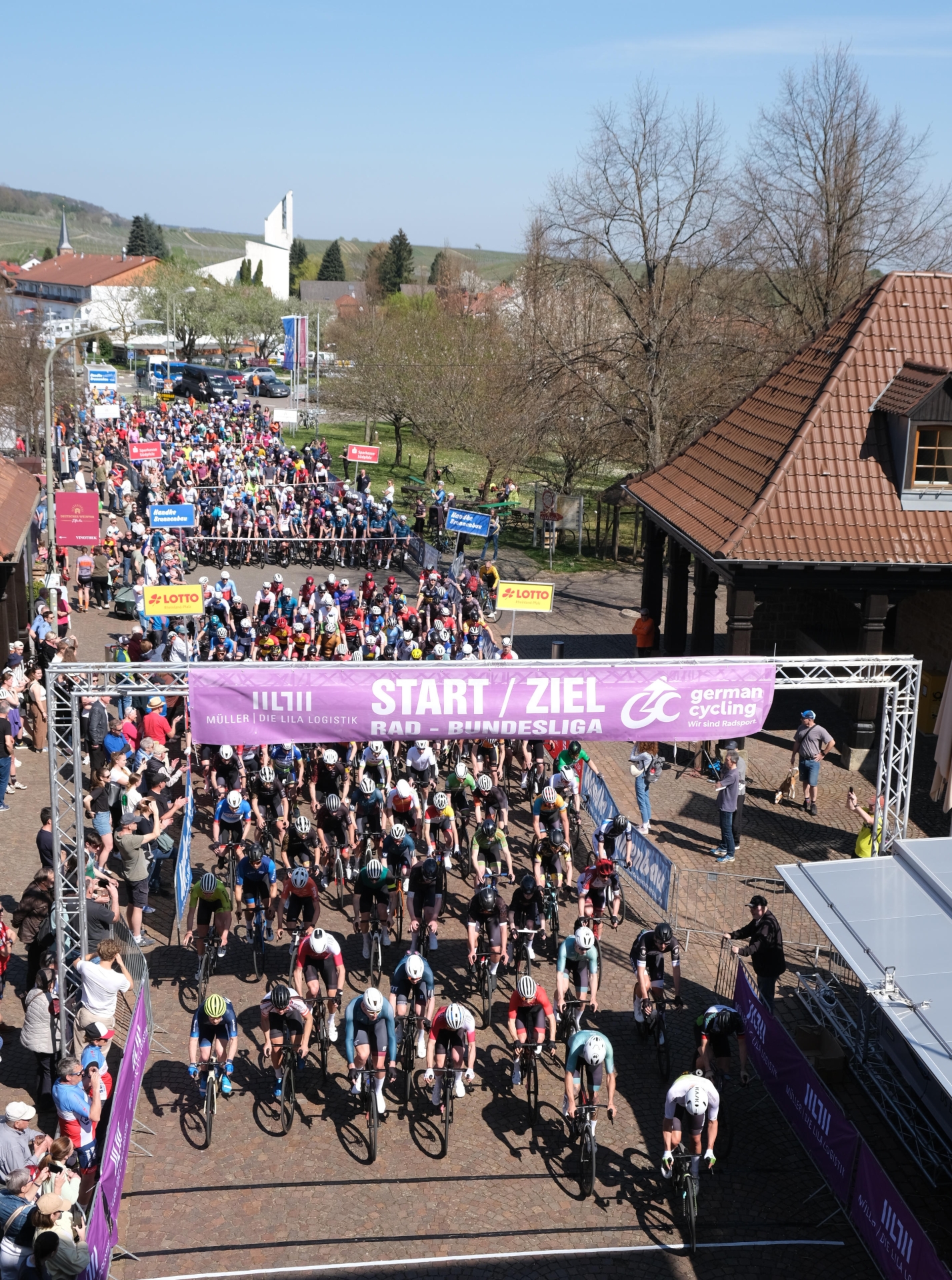 Blick von oben auf das große Starterfeld beim Großen Preis der Südlichen Weinstraße. Zahlreiche Radrennfahrer starten unter dem violetten Start- und Zielbanner der Rad-Bundesliga. Viele Menschen verfolgen das Rennen am Streckenrand.