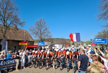 Kurz vor Beginn des Bundesligarennens, das Landrat Dietmar Seefeldt (rechts, mit französischer Flagge) und Wissembourgs Bürgermeisterin Sandra Fischer-Junck (links, mit deutscher Flagge) eröffneten. Foto: KV SÜW Radrennfahrer in orangefarbenen Trikots stehen am Start beim Großen Preis der Südlichen Weinstraße. Zahlreiche Zuschauerinnen und Zuschauer säumen den Startbereich und schwenken Deutschland- und Frankreichflaggen.