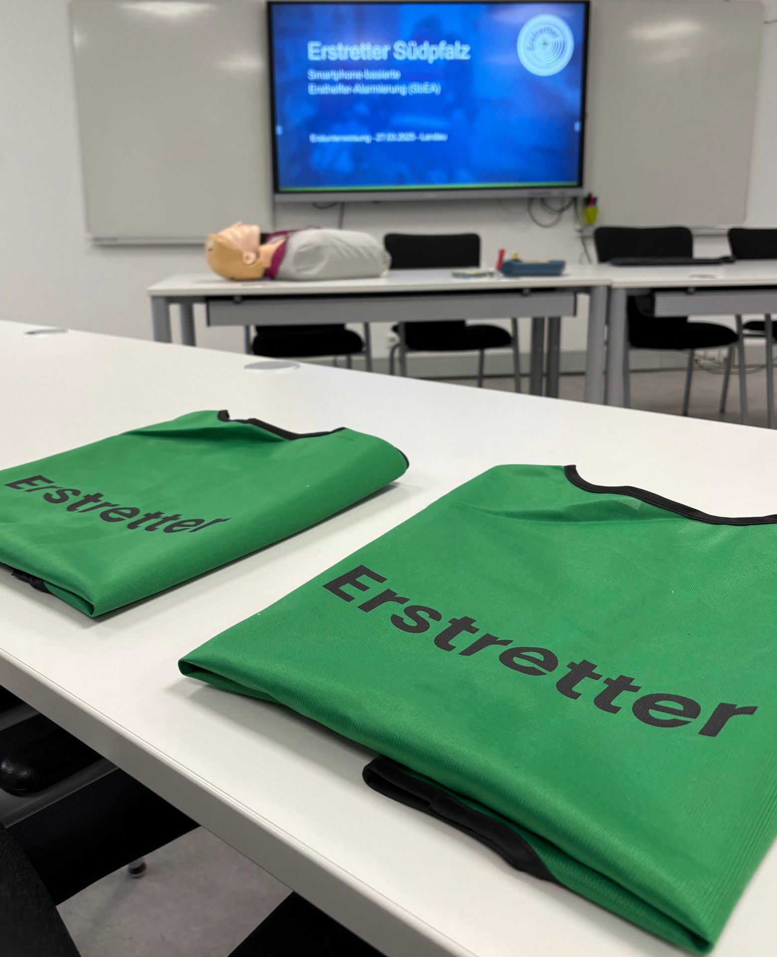 Grüne Westen mit der Aufschrift „Erstretter“ liegen auf einem Tisch in einem Schulungsraum. Im Hintergrund steht eine Übungspuppe auf einem Tisch vor einem Bildschirm, auf dem die Präsentation „Erstretter Südpfalz – Smartphone-basierte Ersthelfer-Alarmierung“ zu sehen ist.