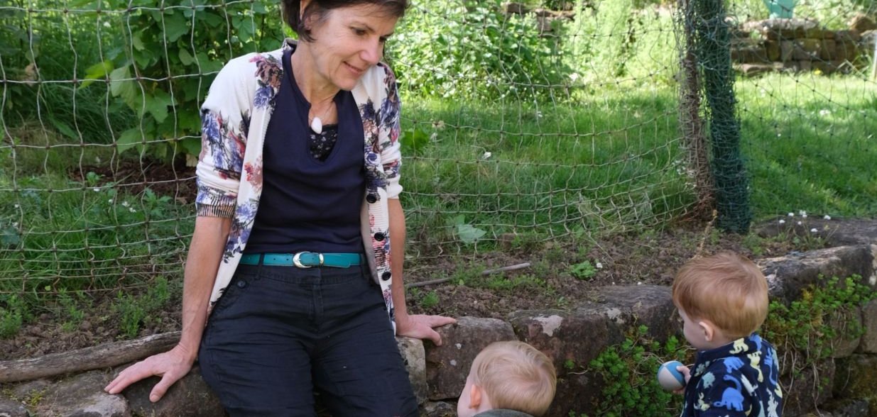 Eine Frau sitzt am Rand einer niedrigen Natursteinmauer in einem grünen Garten und blickt freundlich auf zwei Kleinkinder, die vor ihr stehen. Die Kinder tragen wetterfeste Kleidung und Gummistiefel, eines von ihnen hält ein Spielzeug. Im Hintergrund sind Gemüsebeete, Blumen und ein Zaun zu sehen. Die Szene wirkt ruhig und naturnah.