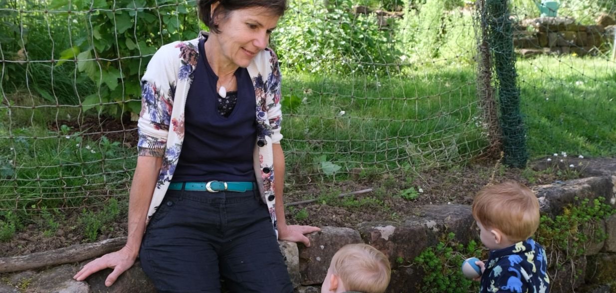 Eine Frau sitzt am Rand einer niedrigen Natursteinmauer in einem grünen Garten und blickt freundlich auf zwei Kleinkinder, die vor ihr stehen. Die Kinder tragen wetterfeste Kleidung und Gummistiefel, eines von ihnen hält ein Spielzeug. Im Hintergrund sind Gemüsebeete, Blumen und ein Zaun zu sehen. Die Szene wirkt ruhig und naturnah.