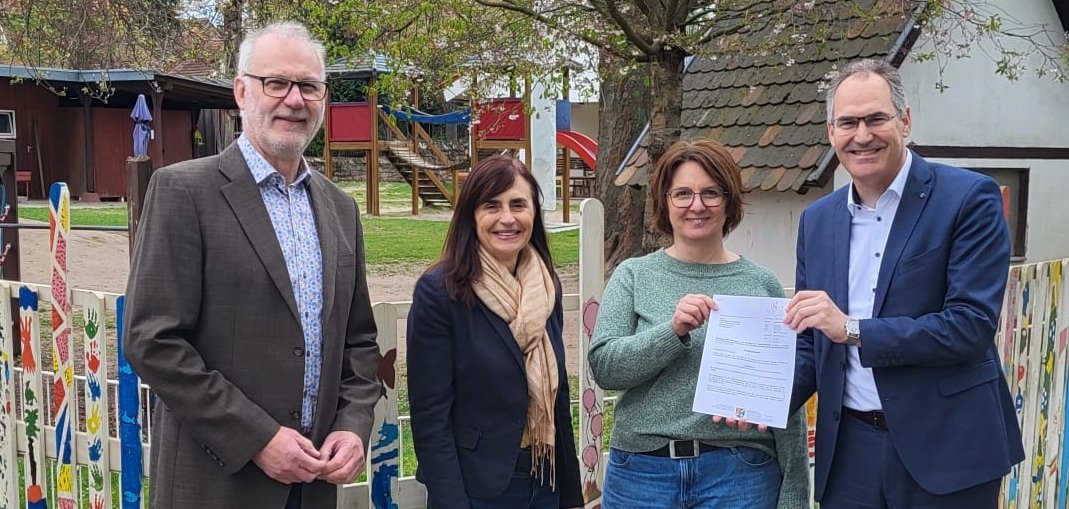 Vier Personen stehen vor einem bunt bemalten Holzzaun auf dem Gelände einer Kindertagesstätte. Ein Mann im Anzug überreicht einer Frau ein offizielles Dokument, beide halten es gemeinsam. Die anderen beiden Personen stehen lächelnd daneben. Im Hintergrund sind ein blühender Baum, ein Spielplatz und ein kleines Gebäude mit Schindeldach zu sehen.