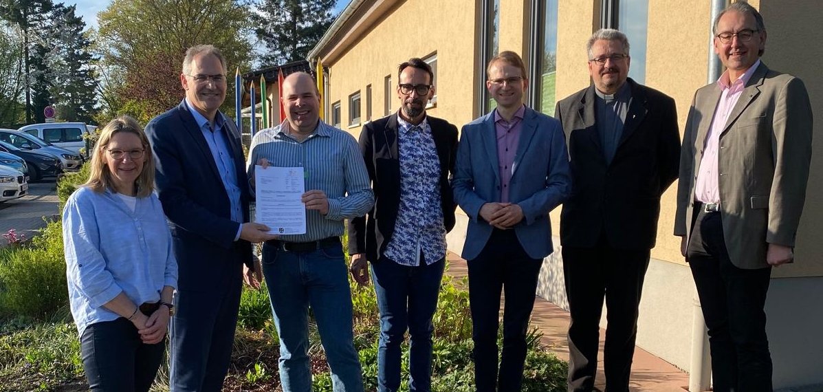 Bei der Bescheidübergabe von links nach rechts: Kita-Leiterin Christine Kiefer, Landrat Dietmar Seefeldt, Dr. Martin Oberhofer, Beigeordneter der Ortsgemeinde, Ortsbürgermeister Christian Kocher, Bürgermeister Daniel Salm, Pfarrer Dr. Achim Dittrich und Erster Kreisbeigeordneter Georg Kern. Die Gruppe steht vor dem Kita-Gebäude in Edesheim.