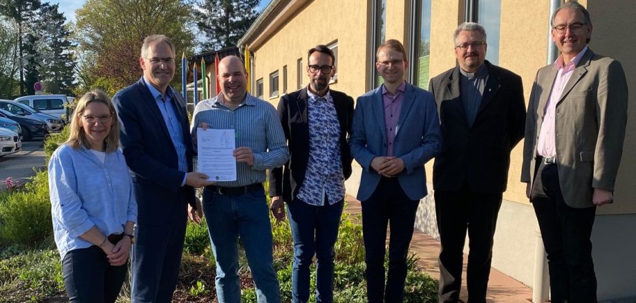 Bei der Bescheidübergabe von links nach rechts: Kita-Leiterin Christine Kiefer, Landrat Dietmar Seefeldt, Dr. Martin Oberhofer, Beigeordneter der Ortsgemeinde, Ortsbürgermeister Christian Kocher, Bürgermeister Daniel Salm, Pfarrer Dr. Achim Dittrich und Erster Kreisbeigeordneter Georg Kern. Die Gruppe steht vor dem Kita-Gebäude in Edesheim.