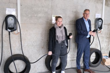 Auch die Ladeinfrastruktur ist da: Agnes Bartmus und Dietmar Seefeldt zeigen die E-Ladesäulen in den Garagen der Kreisverwaltung. Foto: KV SÜW Zwei Personen stehen in einer Garage vor einer Betonwand neben zwei Wandladestationen für E-Fahrzeuge. Beide halten die Ladekabel in der Hand, Reifen dienen als Kabelhalterungen.