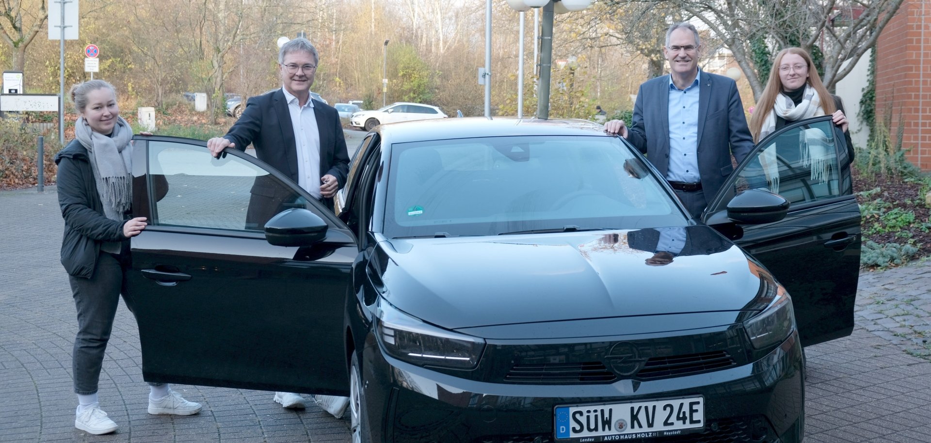 Vier Personen stehen vor einem schwarzen Elektroauto der Kreisverwaltung Südliche Weinstraße mit geöffneten Vordertüren. Das Fahrzeug trägt ein Kennzeichen mit der Aufschrift „SÜW KV 24E“. Im Hintergrund sind Bäume und Verwaltungsgebäude zu sehen.