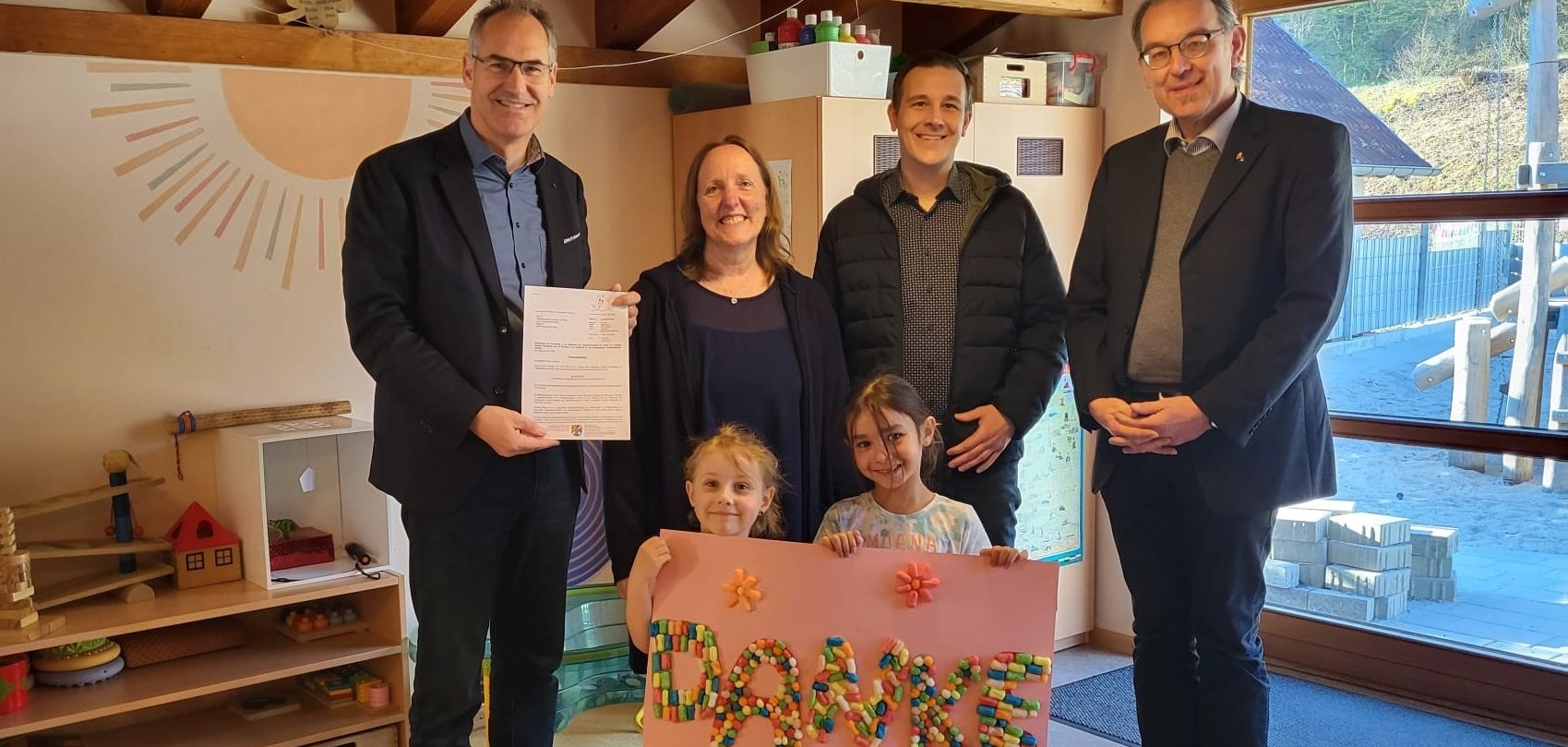 In einem freundlich gestalteten Kita-Raum stehen vier Erwachsene und zwei Kinder. Ein Mann hält ein offizielles Dokument in der Hand, während die beiden Kinder ein großes, bunt gestaltetes Plakat mit der Aufschrift „DANKE“ präsentieren. Im Hintergrund sind Spielzeugregale, Schränke mit Materialien und ein großes Fenster mit Blick auf den Außenbereich der Kita zu sehen.