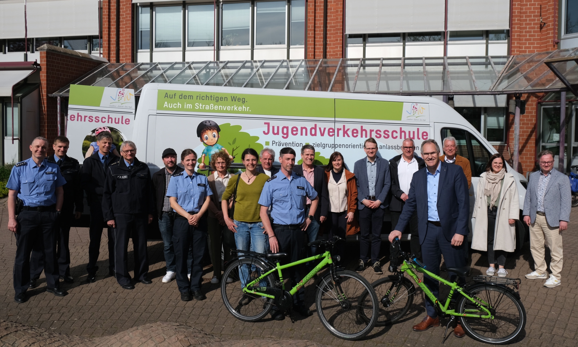 Gruppenbild mit zahlreichen Personen, darunter die uniformierten Verkehrssicherheitsberaterinnen und -berater, die vor dem neuen Fahrzeug der Jugendverkehrsschule stehen, das vor dem Eingang des Kreishauses parkt.
