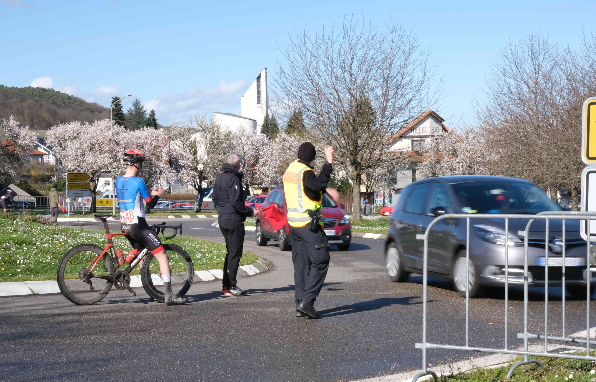 Ein Ordner in Warnweste regelt den Verkehr an einem Kreisverkehr, während ein Radrennfahrer mit schlammverschmutztem Fahrrad am Straßenrand steht. Ein weiteres Teammitglied beobachtet die Situation. Im Hintergrund blühende Bäume, Gebäude und vorbeifahrende Autos.