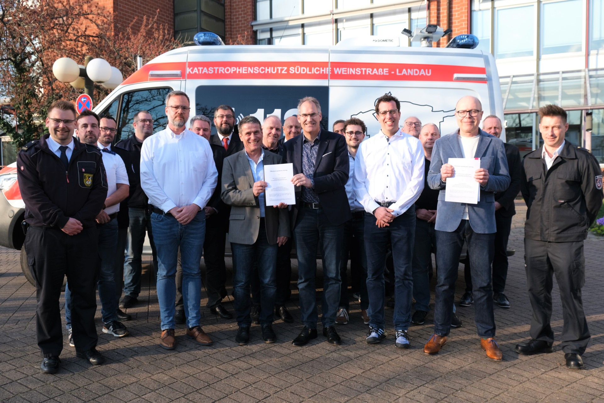 Eine Gruppe von Männern steht vor einem Einsatzfahrzeug mit der Aufschrift „Katastrophenschutz Südliche Weinstraße – Landau“. Zwei Männer in der Mitte halten ein unterzeichnetes Dokument und lächeln in die Kamera. Einige der Anwesenden tragen Uniformen oder Hemden mit Abzeichen, andere sind in Zivilkleidung. Im Hintergrund ist ein modernes Gebäude mit großen Glasfenstern zu sehen.
