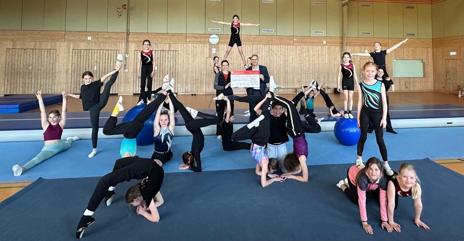 Kinder und Jugendliche einer Turngruppe zeigen gemeinsam verschiedene akrobatische Figuren in einer Sporthalle. In der Mitte des Bildes überreichen zwei Erwachsene einen großen symbolischen Spendenscheck.