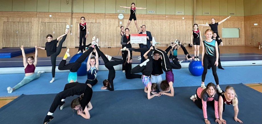 Kinder und Jugendliche einer Turngruppe zeigen gemeinsam verschiedene akrobatische Figuren in einer Sporthalle. In der Mitte des Bildes überreichen zwei Erwachsene einen großen symbolischen Spendenscheck.