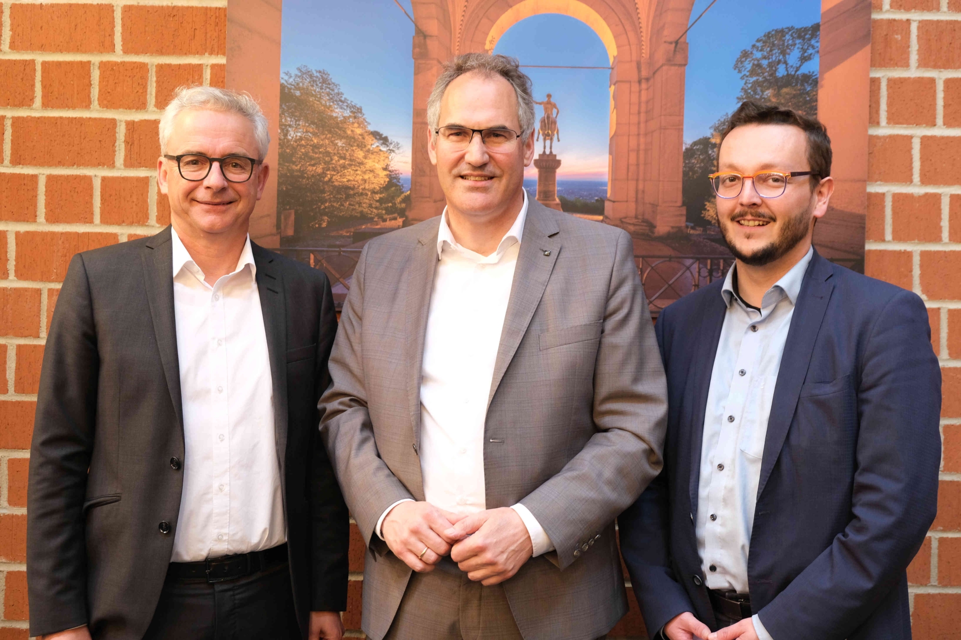 Christoph Schnaudigel, Landrat des Landkreises Karlsruhe und Vizepräsident des Eurodistrikts PAMINA, Dietmar Seefeldt, Landrat des Landkreises Südliche Weinstraße und Präsident des Eurodistrikts PAMINA, und Victor Vogt, Conseiller d`Alsace und Vize-Präsidenten des Eurodistrikts PAMINA, (von links) im Kreishaus SÜW. Sie stehen vor einer Fotografie des Friedensdenkmals Edenkoben im Landkreis SÜW, das Ende des 19. Jahrhunderts als Denkmal für den Sieg im Deutsch-Französischen Krieg errichtet wurde und heute als wichtiges Mahnmal für den Frieden mit den französischen Nachbarn gilt.