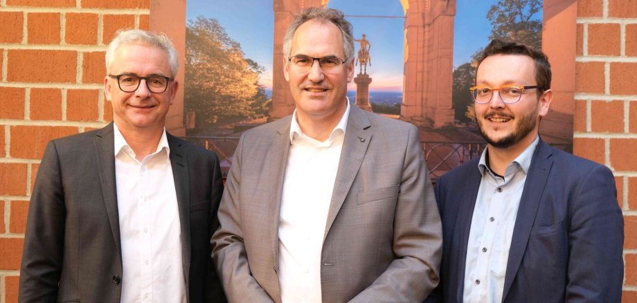 Christoph Schnaudigel, Landrat des Landkreises Karlsruhe und Vizepräsident des Eurodistrikts PAMINA, Dietmar Seefeldt, Landrat des Landkreises Südliche Weinstraße und Präsident des Eurodistrikts PAMINA, und Victor Vogt, Conseiller d`Alsace und Vize-Präsidenten des Eurodistrikts PAMINA, (von links) im Kreishaus SÜW. Sie stehen vor einer Fotografie des Friedensdenkmals Edenkoben im Landkreis SÜW, das Ende des 19. Jahrhunderts als Denkmal für den Sieg im Deutsch-Französischen Krieg errichtet wurde und heute als wichtiges Mahnmal für den Frieden mit den französischen Nachbarn gilt.