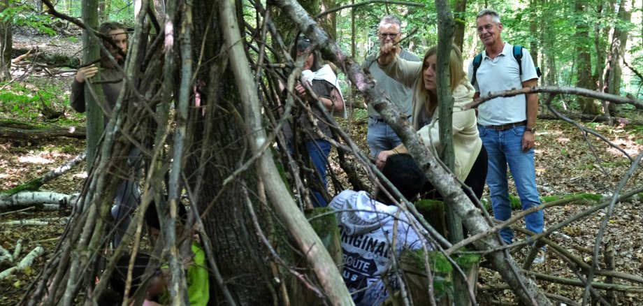 Nahaufnahme eines Tipis aus Ästen und Naturmaterialien in einem Waldstück. Einige erwachsene begutachten das Tipi.
