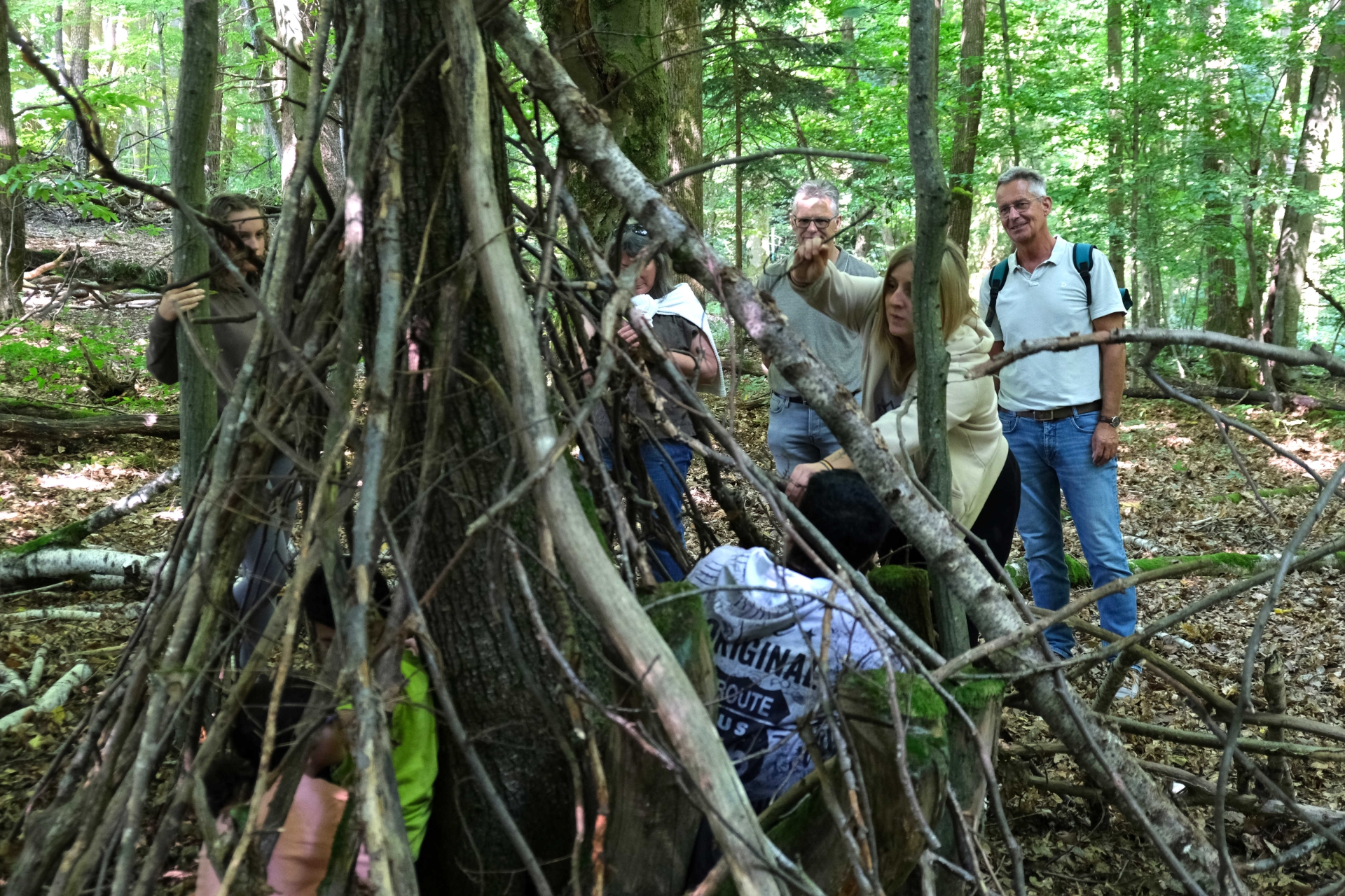 Nahaufnahme eines Tipis aus Ästen und Naturmaterialien in einem Waldstück. Einige erwachsene begutachten das Tipi.