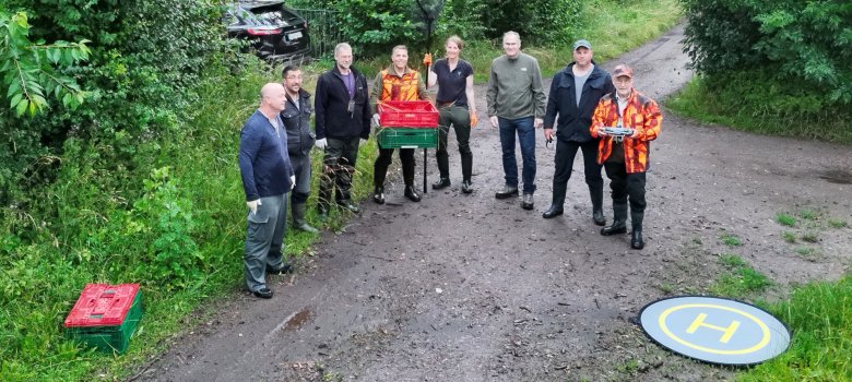 Mehrere Männer stehen auf einem Waldweg nebeneinander, einer hält ein Fernsteuergerät, vor ihnen liegt eine Drohne.