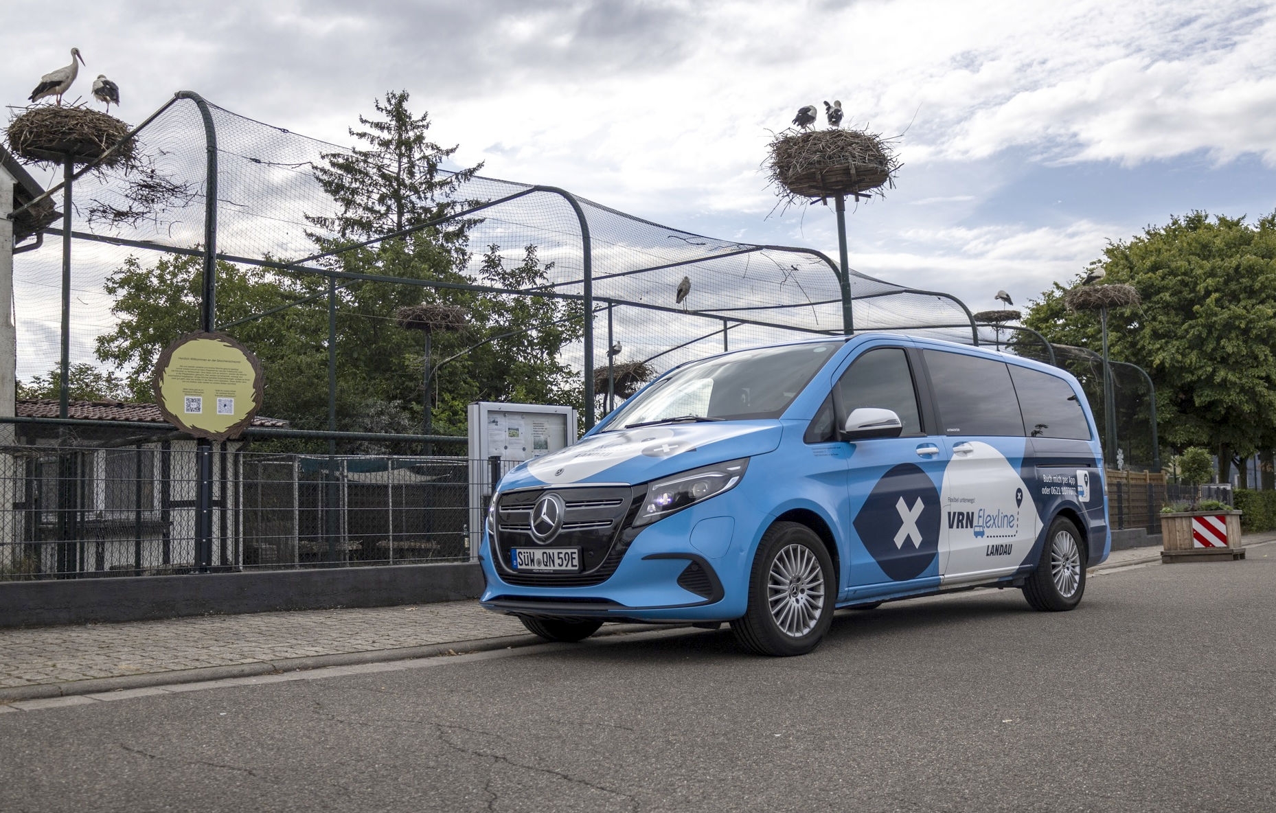 Ein Kleinbus steht vor einem Zaun, dahinter mehrere Nester mit einem Weißstorch.