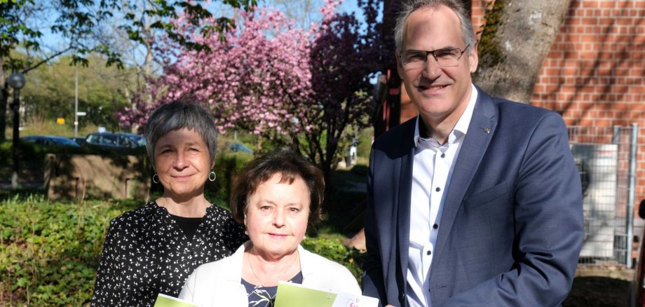 Bettina Forster (links), Brigitte Kültzer und Landrat Dietmar Seefeldt stehen zusammen vor dem Kreishaus in Landau. Im Hintergrund sind unscharf eine Backsteinwand und ein Baum mit rosa Blüten zu sehen. Alle drei lächeln in die Kamera.