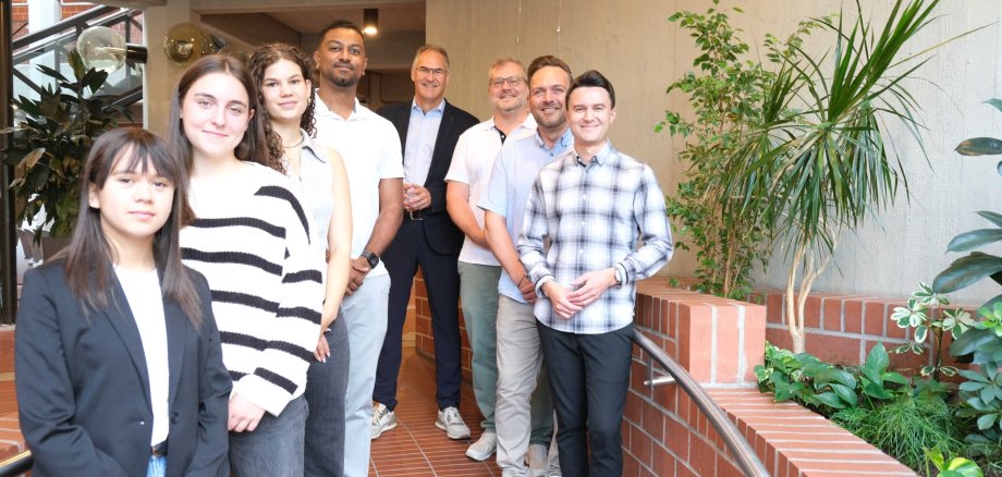 Eine Gruppe von acht Personen steht im Kreishaus SÜW in einer hellen Flur-Passage mit Pflanzen und Backsteinwänden. In der vorderen Reihe stehen vier junge Menschen, dahinter vier Männer in unterschiedlichen Outfits, darunter Landrat Dietmar Seefeldt in Anzug. Die Personen lächeln in die Kamera.