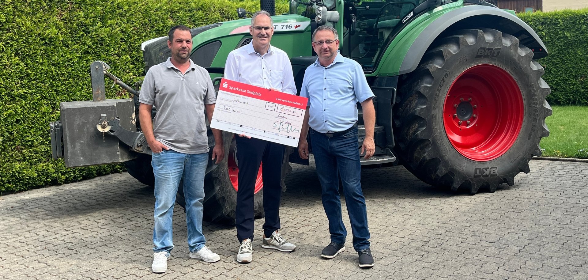 Drei Männer stehen vor einem großen grünen Traktor. Zwei von ihnen tragen Freizeitkleidung, einer ein weißes Hemd. Gemeinsam halten sie einen großen symbolischen Scheck der Sparkasse Südpfalz in der Hand.