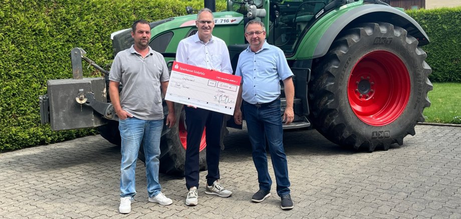 Landrat Dietmar Seefeldt (Mitte) übergibt den Scheck der Stiftung der Sparkasse Südpfalz an die „Red Farmer“-Gründer Ingo Pfalzgraf (links) und Thomas Knecht. Foto: Red Farmer Drei Männer stehen vor einem großen grünen Traktor. Zwei von ihnen tragen Freizeitkleidung, einer ein weißes Hemd. Gemeinsam halten sie einen großen symbolischen Scheck der Sparkasse Südpfalz in der Hand.