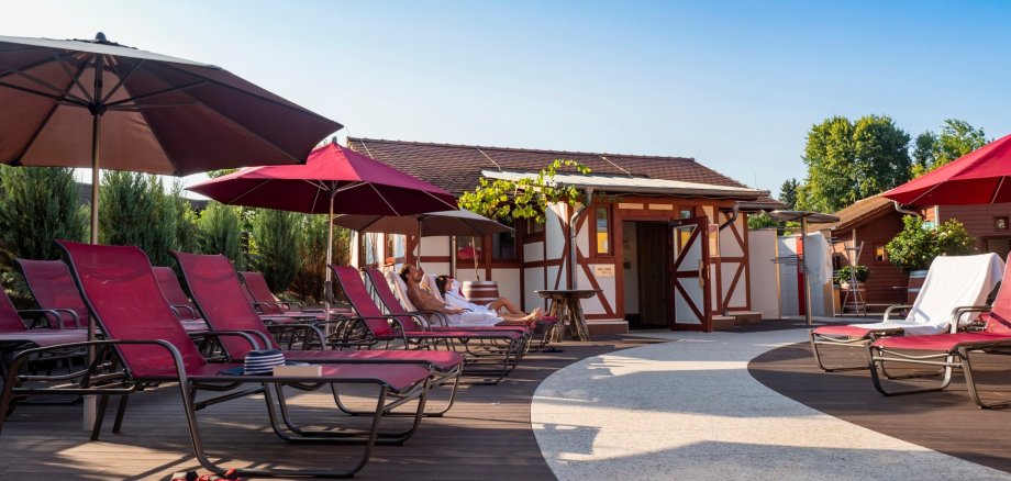 Eine Saunalandschaft in einem Dachgarten unter blauem Himmel: Ein geschwungener Weg aus hellen Steinplatten führt zwischen mit WPC-Dielen ausgelegten Terrassenplätzen hindurch, auf denen rote Liegen unter ebenfalls roten Sonnenschirmen stehen. Auf zwei dieser Sonnenliegen haben ein Mann und eine Frau im Saunatuch platzgenommen. Im Hintergrund ist ein kleines Gebäude im Fachwerkstil zu sehen, das einen Teil des Saunaangebots beherbergt.