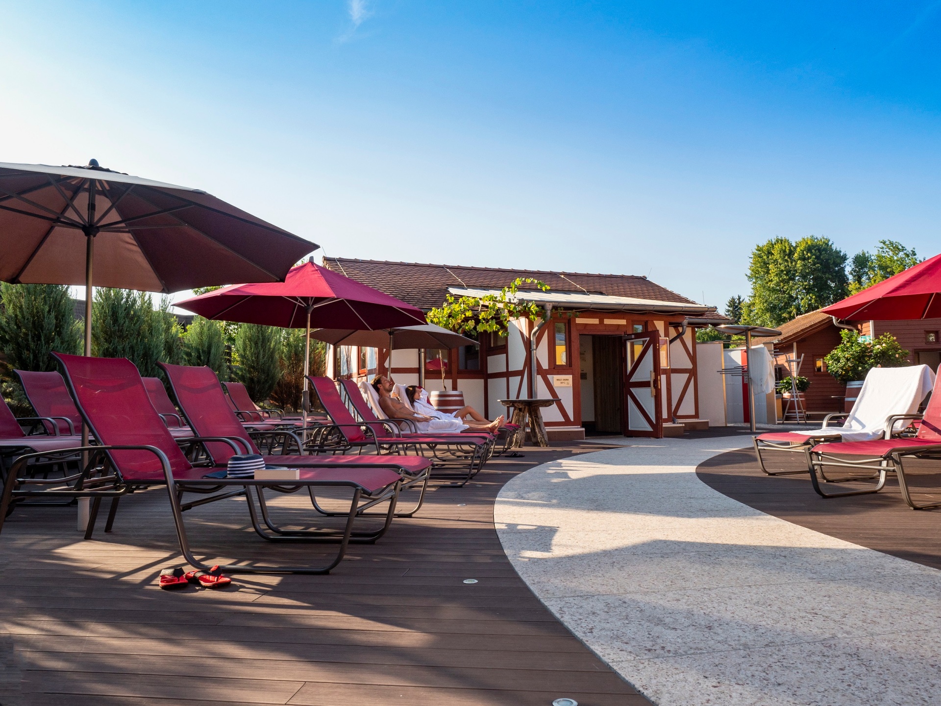 Eine Saunalandschaft in einem Dachgarten unter blauem Himmel: Ein geschwungener Weg aus hellen Steinplatten führt zwischen mit WPC-Dielen ausgelegten Terrassenplätzen hindurch, auf denen rote Liegen unter ebenfalls roten Sonnenschirmen stehen. Auf zwei dieser Sonnenliegen haben ein Mann und eine Frau im Saunatuch platzgenommen. Im Hintergrund ist ein kleines Gebäude im Fachwerkstil zu sehen, das einen Teil des Saunaangebots beherbergt.