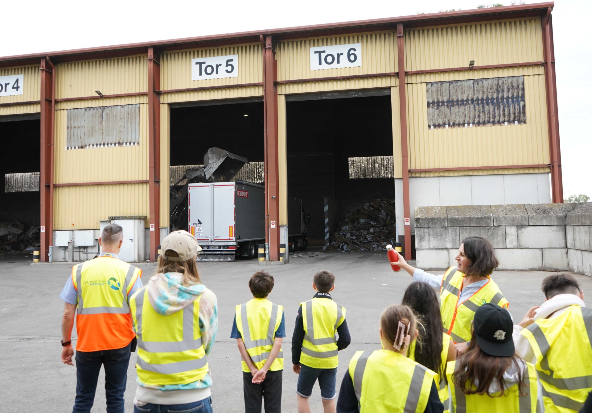 Eine Gruppe blickt auf eine Halle mit hohen Toren, in einem Tor steht ein LKW, der gerade von einem Radlader beladen wird.