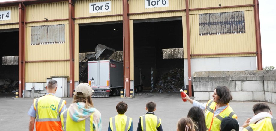 Eine Gruppe blickt auf eine Halle mit hohen Toren, in einem Tor steht ein LKW, der gerade von einem Radlader beladen wird.