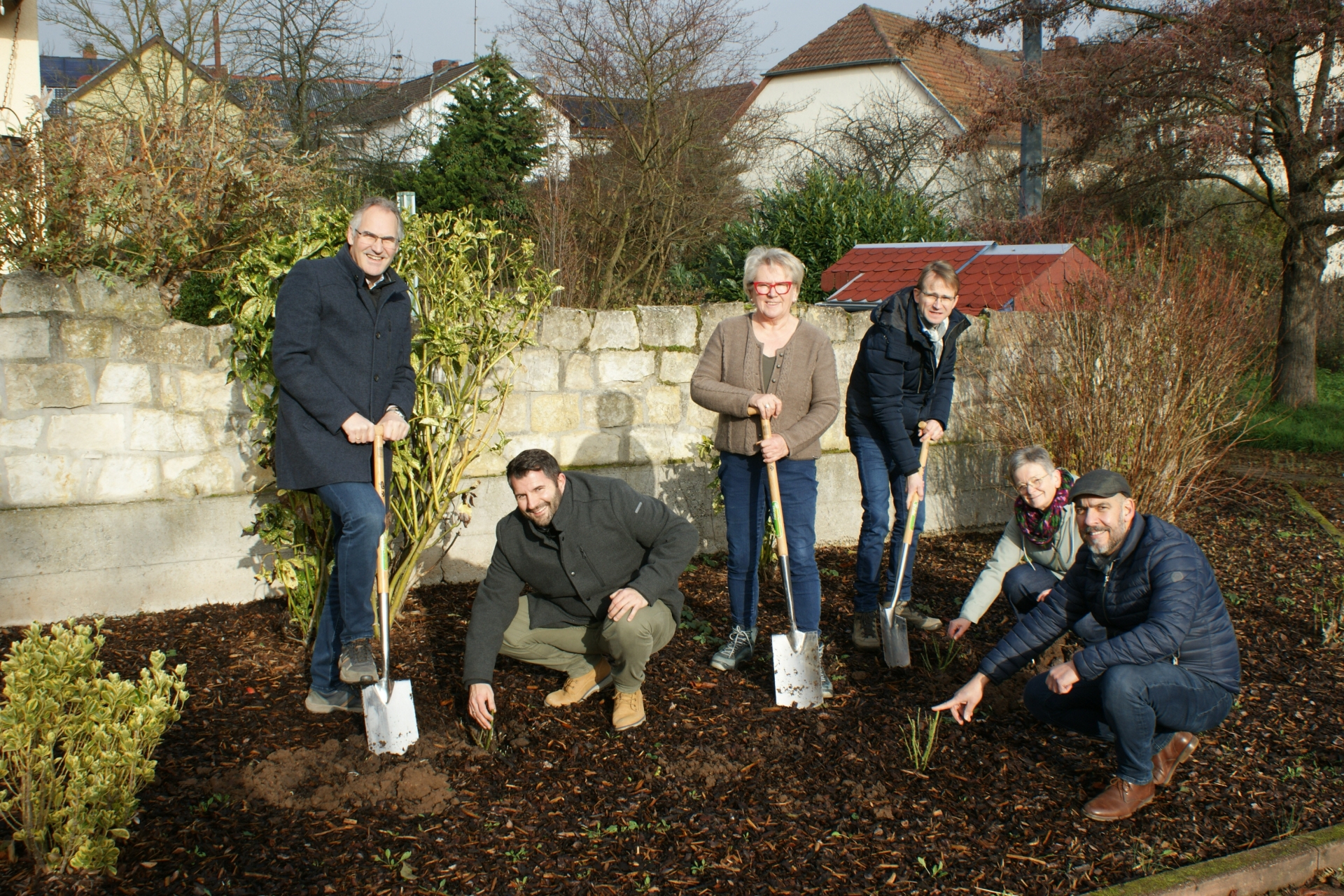 Mehrere Personen, teilweise mit Spaten in der Hand, stehen neben frisch gepflanzten Rosen in einem Blumenbeet und lächeln in die Kamera.