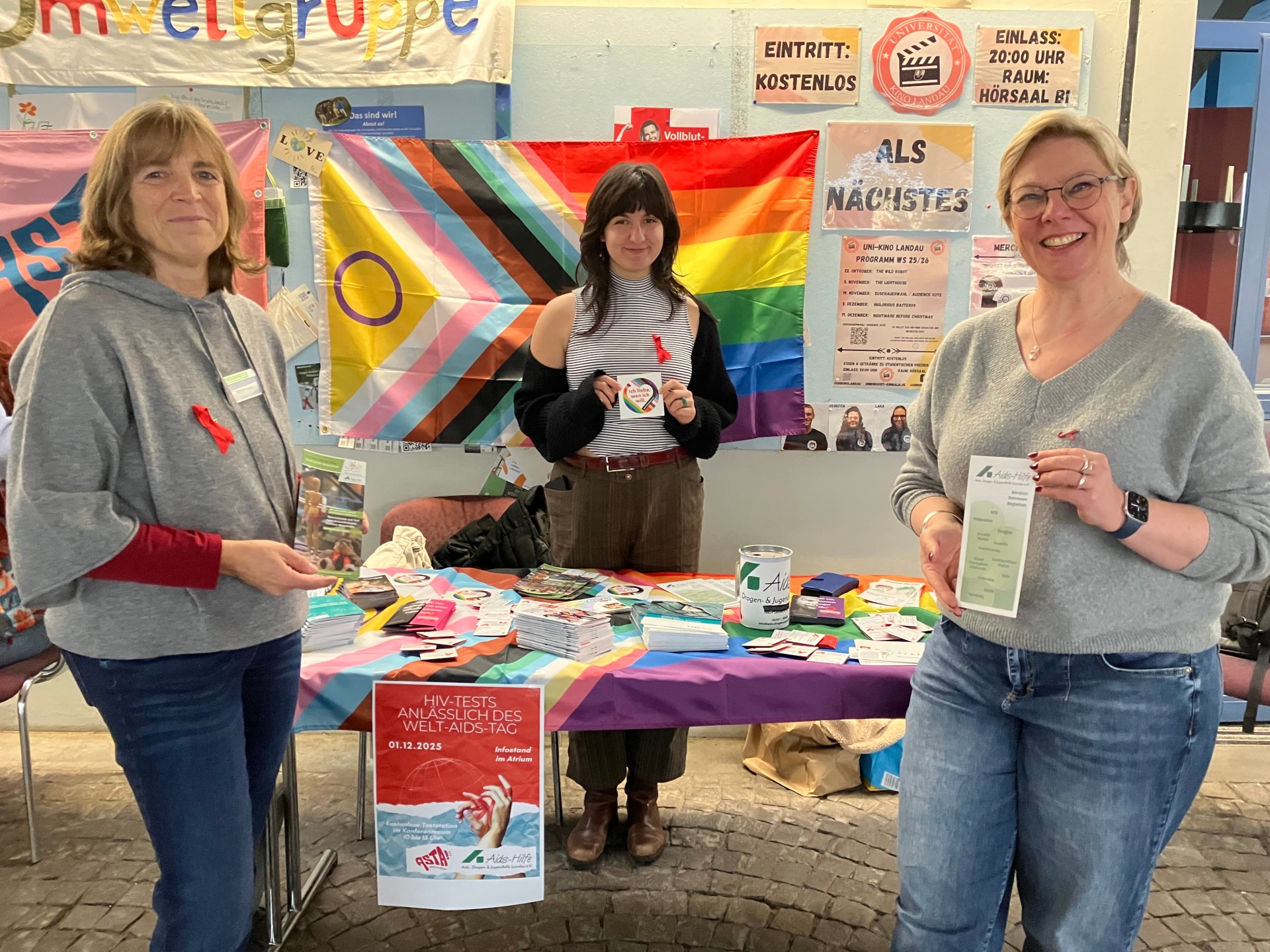 Drei Personen stehen vor einer bunten Wand und einem Infotisch im Atrium der Landauer Uni, sie tragen eine rote Schleife angeheftet.