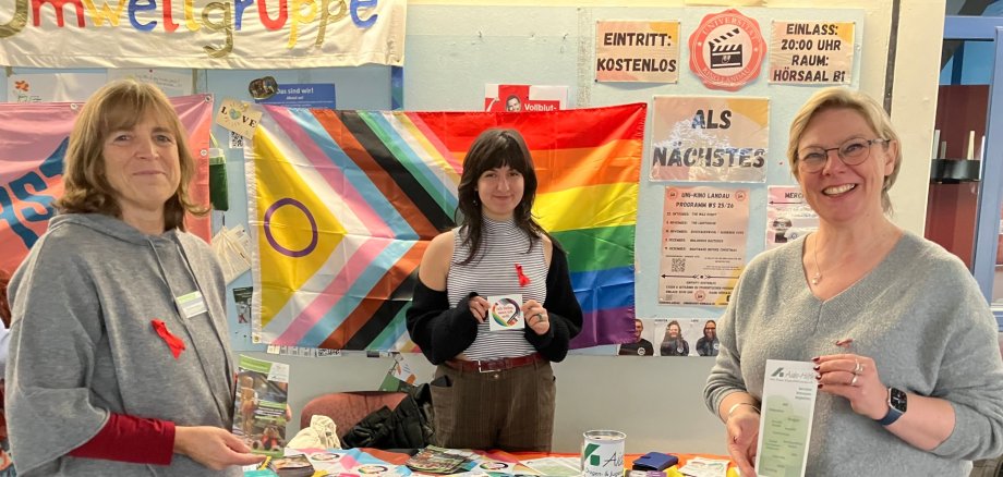 Drei Personen stehen vor einer bunten Wand und einem Infotisch im Atrium der Landauer Uni, sie tragen eine rote Schleife angeheftet.