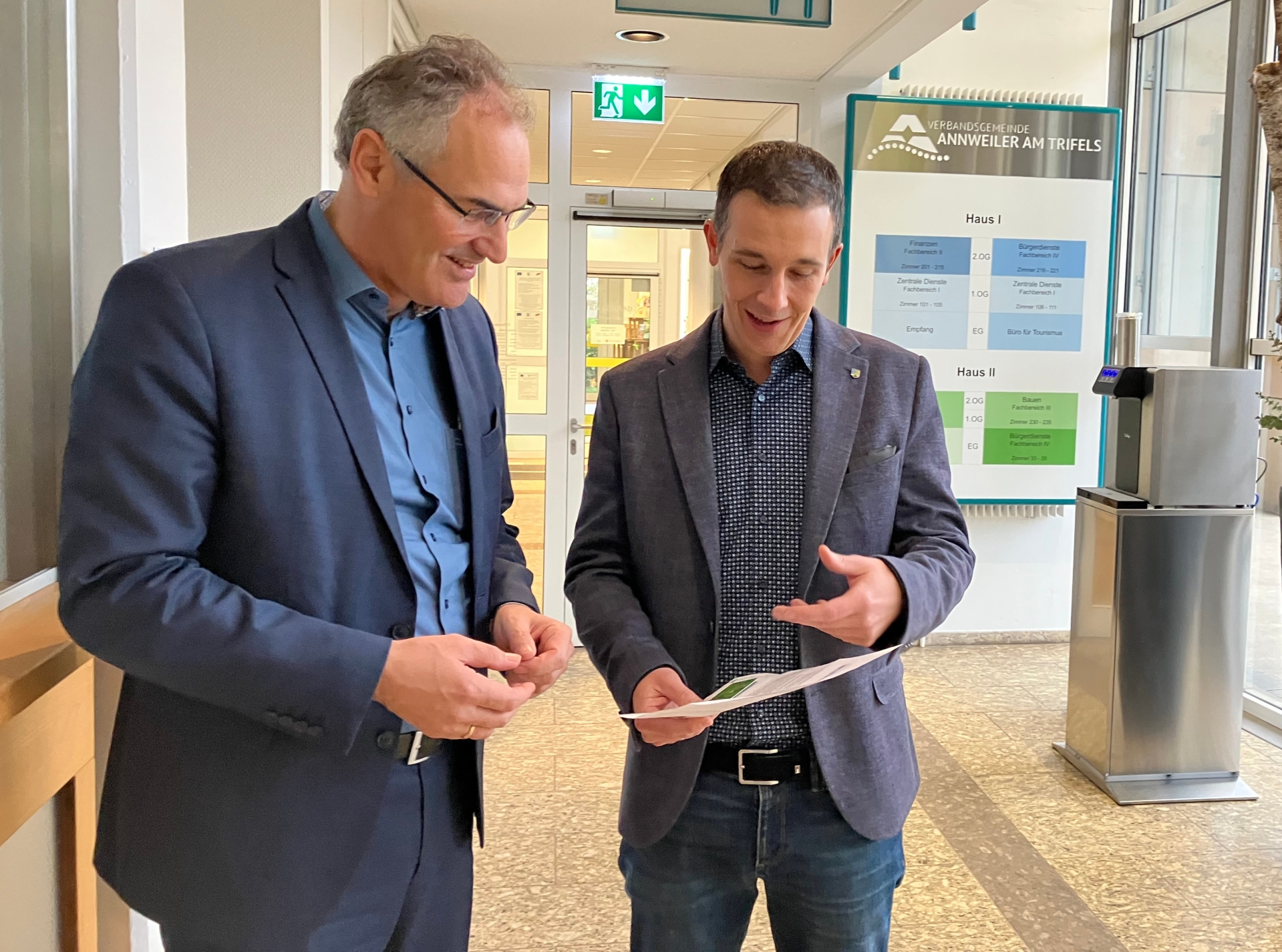 Landrat Dietmar Seefeldt und Bürgermeister Christian Burkhart stehen in einem Flur und unterhalten sich über ein Dokument, das sie gemeinsam betrachten. Im Hintergrund ist ein Schild mit dem Logo der Verbandsgemeinde Annweiler am Trifels zu sehen.