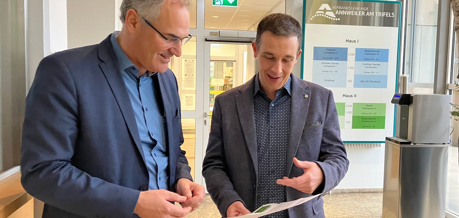 Landrat Dietmar Seefeldt und Bürgermeister Christian Burkhart stehen in einem Flur und unterhalten sich über ein Dokument, das sie gemeinsam betrachten. Im Hintergrund ist ein Schild mit dem Logo der Verbandsgemeinde Annweiler am Trifels zu sehen.