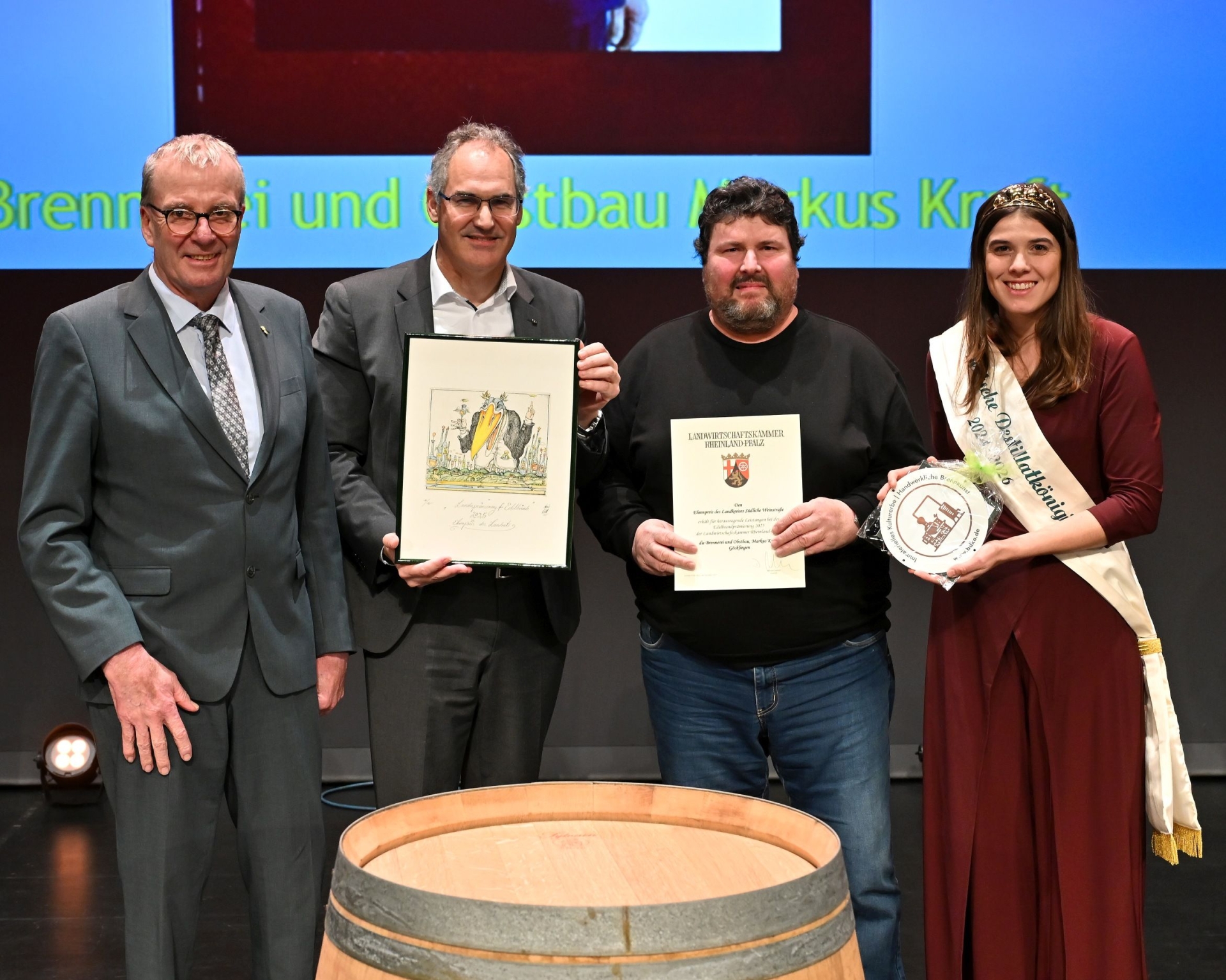 Drei Männer und eine Frau stehen um ein Holzfass, einer hält eine Urkunde, einer ein gerahmtes Bild. Die Frau trägt eine Schärpe mit der Aufschrift "Deutsche Destillatkönigin."