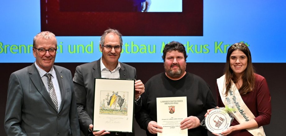 Drei Männer und eine Frau stehen um ein Holzfass, einer hält eine Urkunde, einer ein gerahmtes Bild. Die Frau trägt eine Schärpe mit der Aufschrift "Deutsche Destillatkönigin."