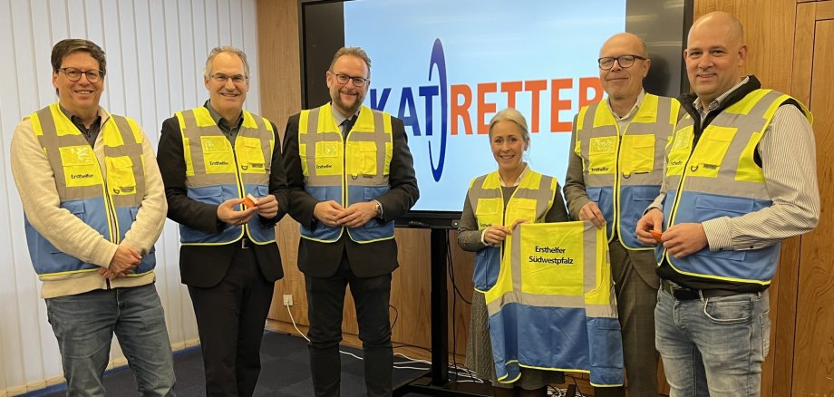 Sechs Personen stehen nebeneinander und lächeln in die Kamera. Sie alle tragen eine gelbe Weste, auf der "Ersthelfer" steht.