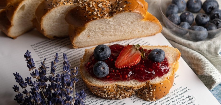 Aufgeschnittenes Früstücksbrot mit Marmelade, Heidelbeeren und einer halben Erdbeere liegt auf einem aufgeschlagenen Buch. Daneben liegt ein kleiner Strauß Lavendel. Neben dem Buch steht eine Schale mit weiteren Heidelbeeren.