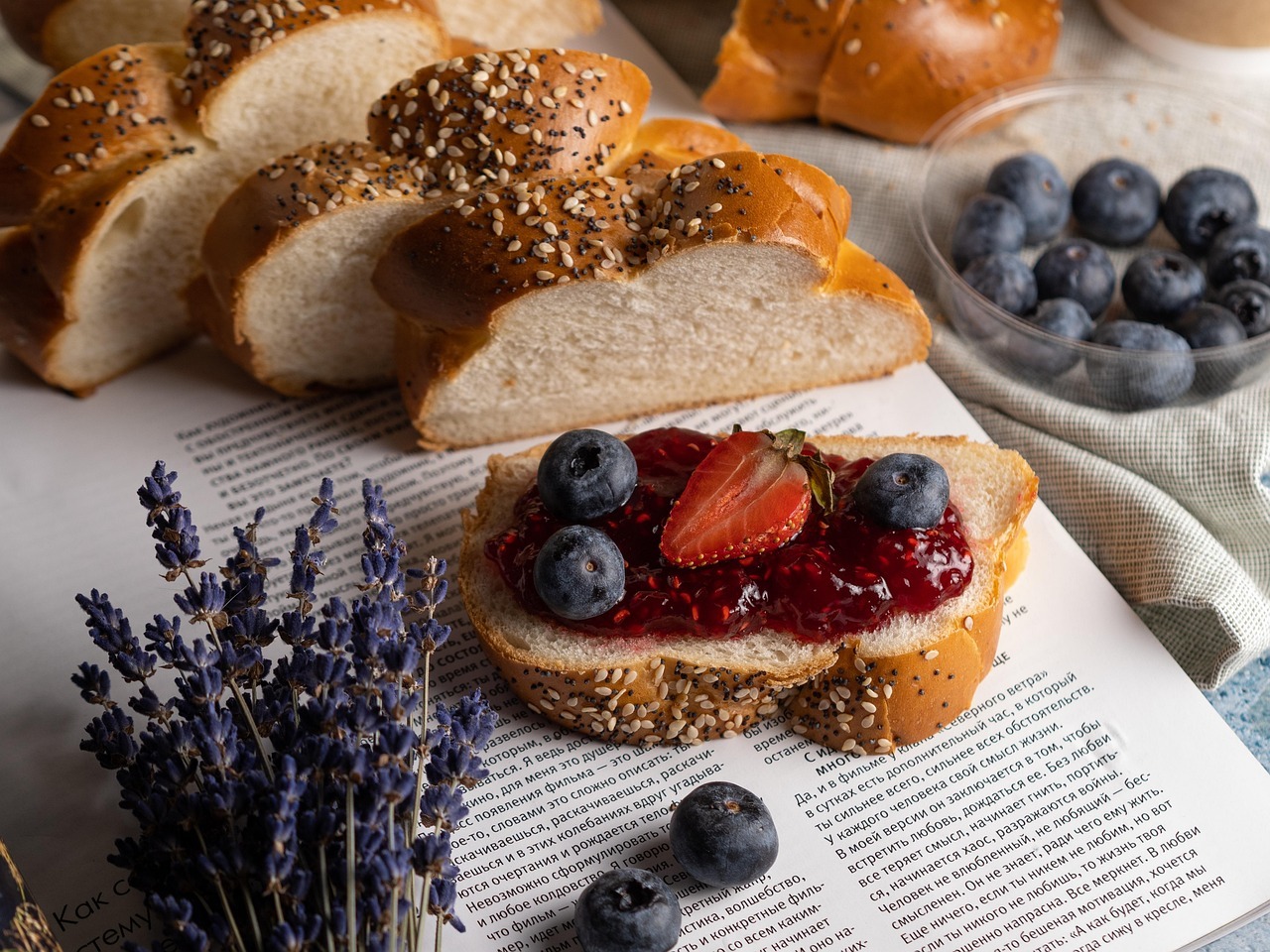 Aufgeschnittenes Früstücksbrot mit Marmelade, Heidelbeeren und einer halben Erdbeere liegt auf einem aufgeschlagenen Buch. Daneben liegt ein kleiner Strauß Lavendel. Neben dem Buch steht eine Schale mit weiteren Heidelbeeren.
