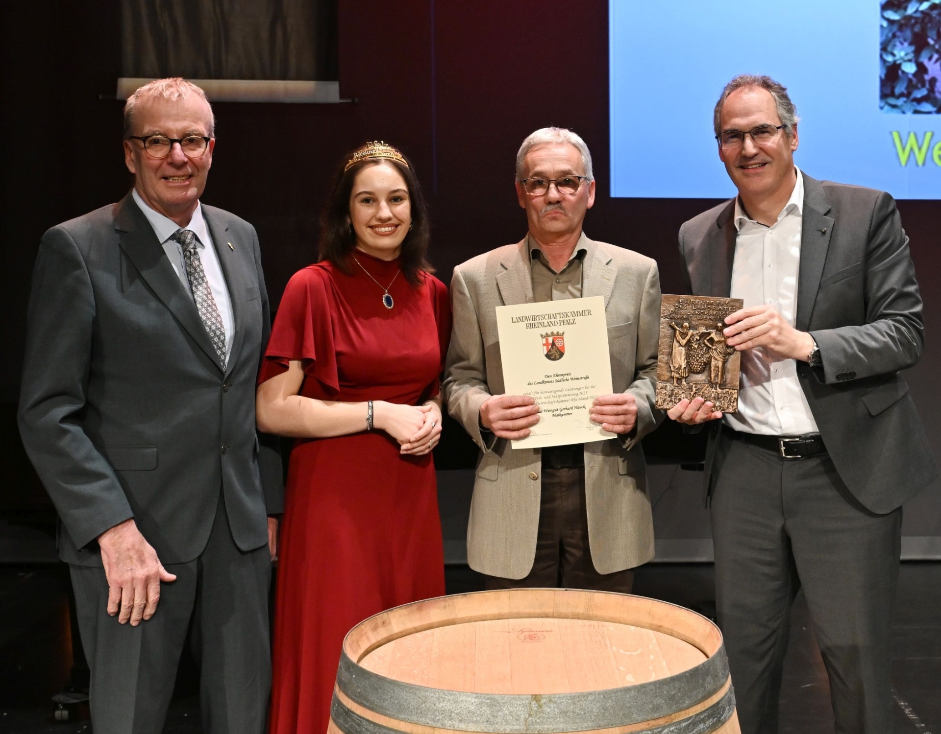 Drei Männer und eine Frau stehen um ein Holzfass. Die Frau trägt ein goldenes Diadem, ein Mann hält eine Urkunde, ein anderer eine bronzene Plakette.